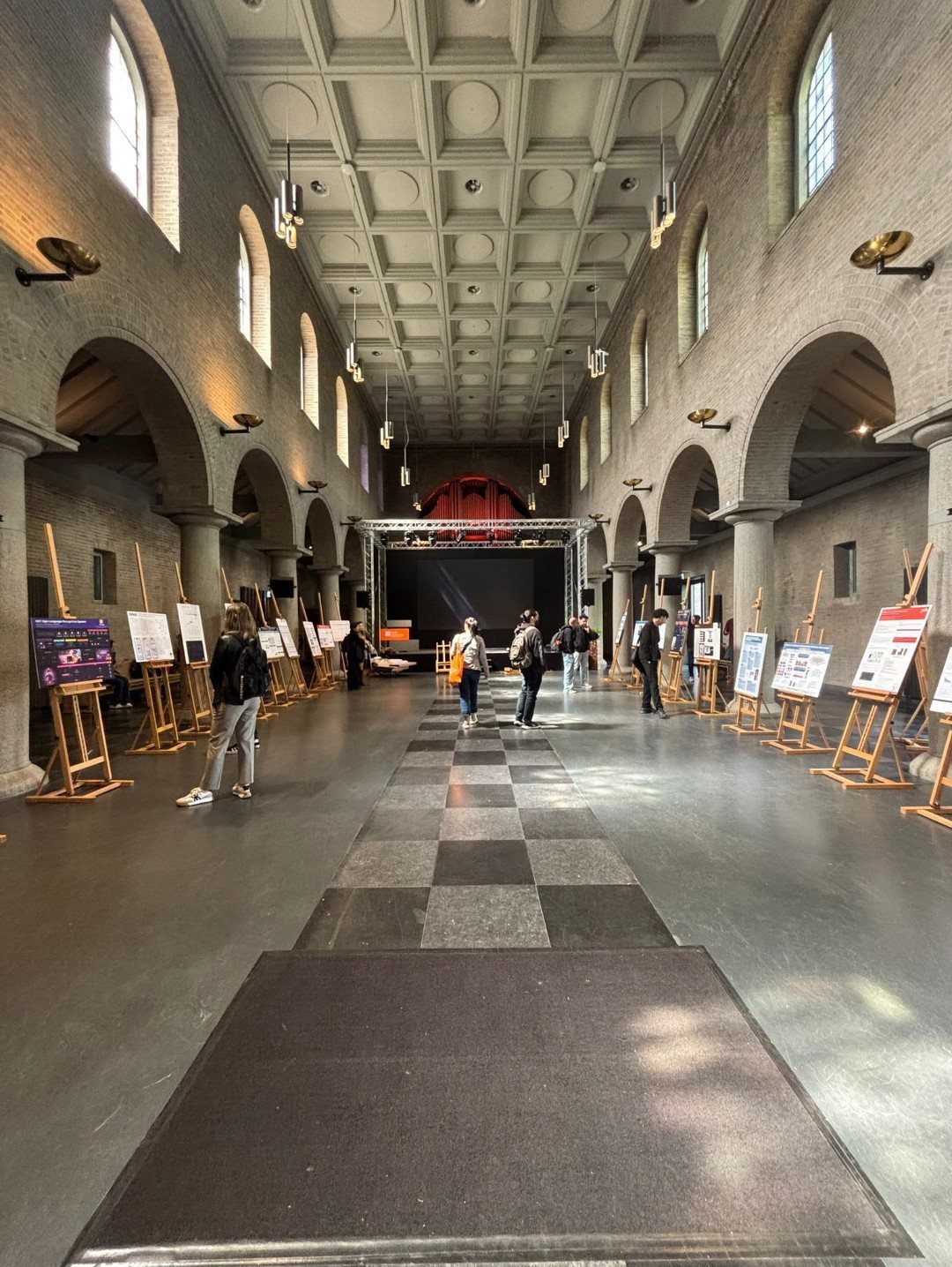 A long brick hall with arches and wooden easels holding posters on both sides, with visitors looking at them