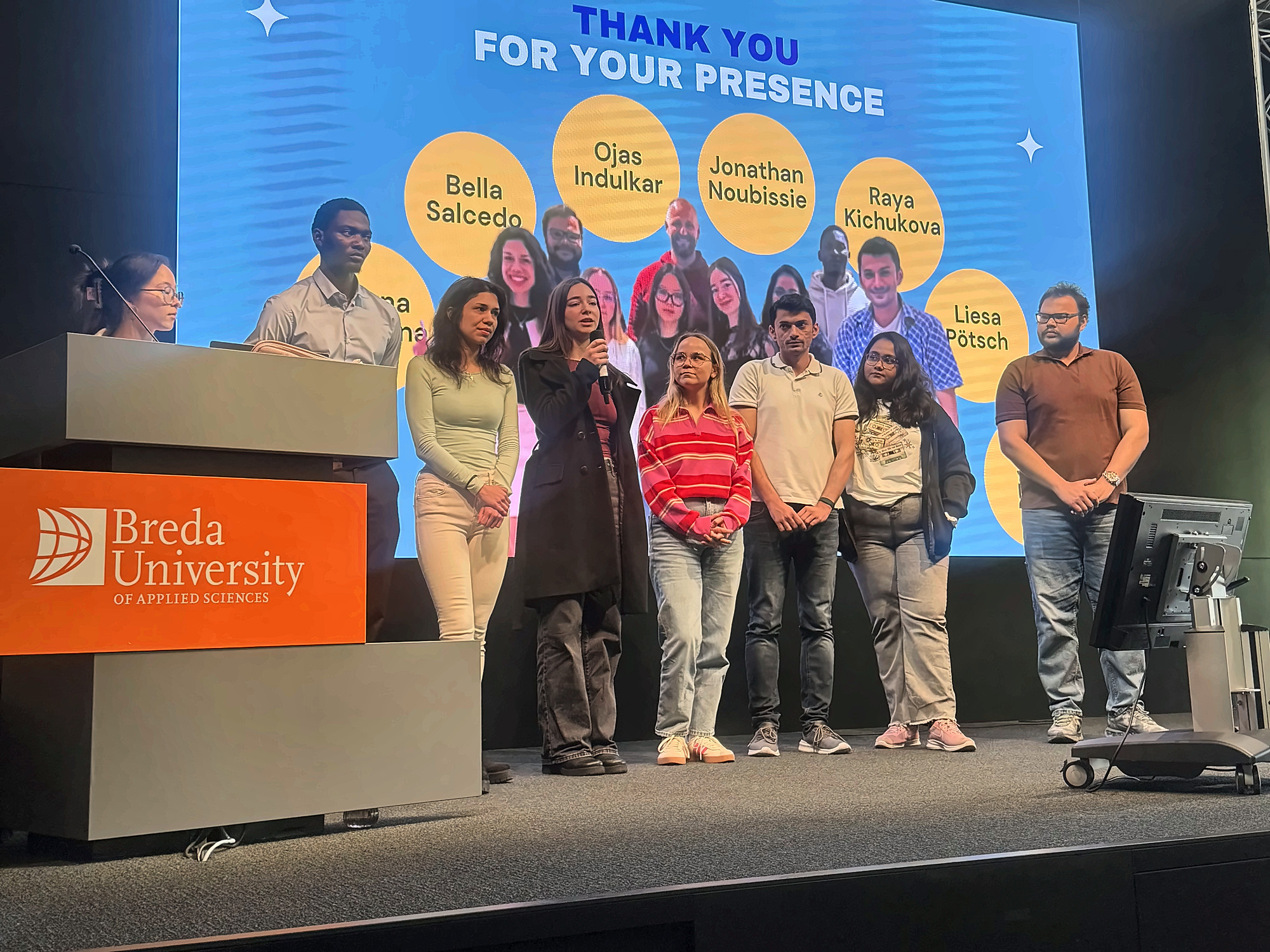 The whole team standing together on the Chapel stage while one member answers a question into a microphone, in front of a slide reading Thank you for your presence